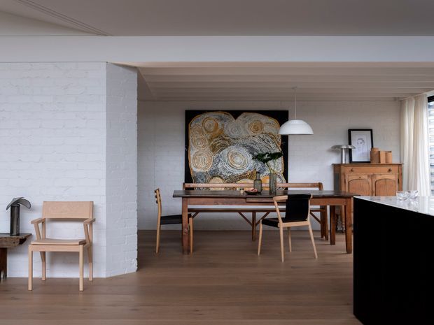 Open-plan dining area with warm timber flooring, a timber dining table and chairs, white-painted brick walls, and a timber-lined ceiling. A large abstract artwork anchors the dining setting, with a sideboard and pendant light adding mid-century character. The foreground includes part of a dark kitchen island or bench.