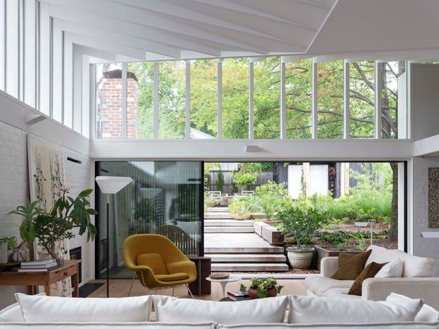 Bright contemporary living room with a white sectional sofa, mustard lounge chair, indoor plants, and artwork. Floor-to-ceiling glazing and a high clerestory window wall open directly onto a landscaped multi-level courtyard garden, creating a strong indoor–outdoor connection. The room has a white painted interior, exposed white ceiling boards, dark-framed glazing, and abundant natural light.