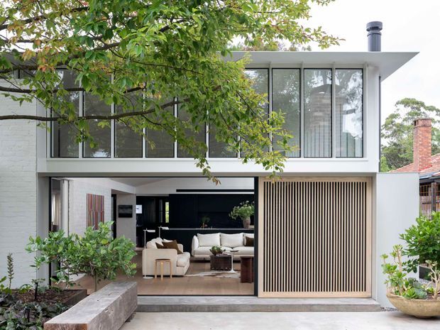 Contemporary two-storey residential facade with a crisp white rendered and brick exterior, broad black-framed upper-level glazing, and a recessed ground-floor living area opening onto the front. A tall vertical timber screen conceals part of the lower facade, while mature planting and a concrete garden planter frame the entry. The flat roof and strong horizontal composition create a clean modern architectural expression.