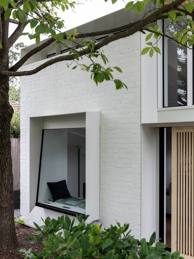 Exterior view of a contemporary white-painted brick house partially screened by mature tree branches and foliage. The facade features deep-set dark-framed windows, a projecting white window surround, a flat roofline, and a sheltered opening with vertical timber screening. A glimpse of an interior window seat is visible through the lower window.