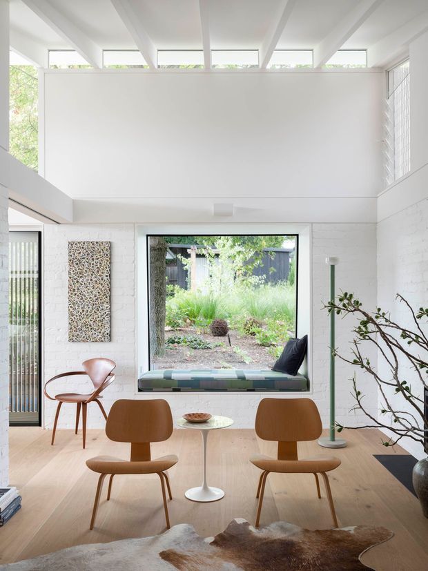 Bright double-height sitting and dining area with white-painted brick walls, exposed white ceiling beams, clerestory windows and large black-framed glazing overlooking a landscaped courtyard. A built-in window seat, sculptural floor lamp, mid-century timber chairs, small round pedestal table and textured wall artwork create a refined mid-century interior.