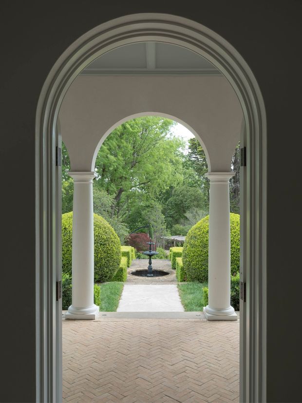A view from inside a tall, arched doorway toward a formal garden courtyard. The architectural opening has layered curved mouldings, white classical columns, and a shallow vaulted or coffered ceiling detail. Outside, clipped hedges, rounded topiary, a central stone path, and a small ornamental fountain create a symmetrical garden composition. The foreground floor is laid in a pale herringbone brick pattern. The image has a refined, traditional character with soft natural light and strong axial symmetry.