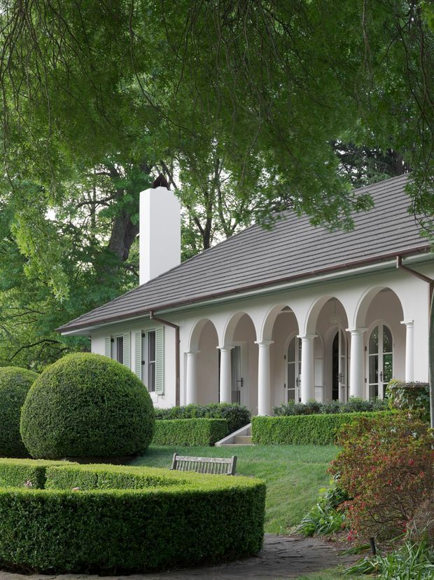 Exterior view of a refined single-storey residence set within mature, heavily landscaped grounds. The building has a long pitched roof with grey tile-like roofing, pale rendered walls, a prominent white chimney, and a covered verandah defined by a repeated series of rounded arches and slender columns. Formal clipped hedges, sculpted shrubs, lawn, and a paved garden path frame the facade.