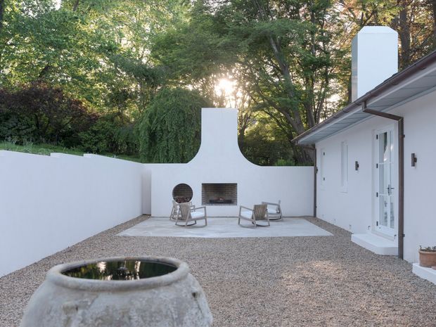 A minimalist white-walled courtyard viewed from a gravel-covered outdoor area. The courtyard features a built-in outdoor fireplace with a sculptural chimney, two light-coloured lounge chairs, a small circular fire feature or opening, and a large weathered ceramic vessel in the foreground. A white building with simple doors and a low sloping roof opens directly onto the courtyard, while mature trees provide a leafy backdrop.