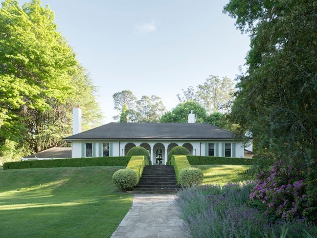 A symmetrical single-storey house viewed from the front, set within a formal landscaped garden. The low, dark hipped roof is supported by a white facade with a central arched colonnade and matching white chimneys. A straight stone-paved path leads through clipped hedges and lawns to the entrance, with mature trees and flowering planting framing the property.