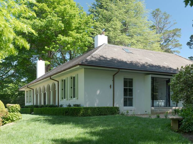 A single-storey, low-pitched hipped-roof house set within a mature landscaped garden. The light-painted exterior features white rendered walls, a tiled roof, traditional multi-pane windows with shutters, chimneys, and a distinctive covered side verandah with repeated rounded arches. A skylight is visible on the roof, and neatly clipped hedges frame the lawn.