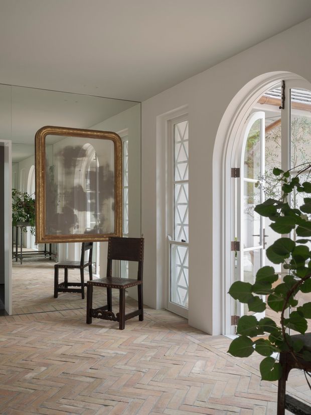 A refined interior hallway or entry space with pale plaster walls, arched white-framed glazing and a narrow geometric-paned door. The floor is laid in a warm patterned brick or terracotta herringbone finish. A large antique-style gilt-framed mirror, dark timber chairs, and a leafy indoor tree add character. Full-height mirrored wall panels visually extend the corridor and reflect the arched openings and furnishings. The space has a bright, warm, layered aesthetic with traditional architectural detailing.