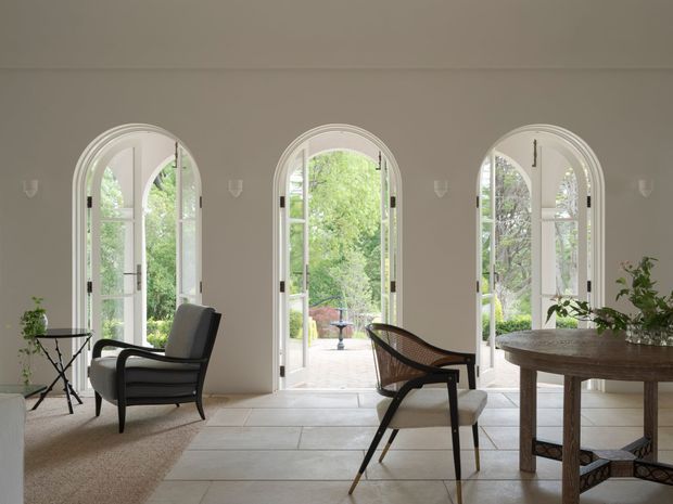Elegant, light-filled interior with three tall arched glazed French doors opening to a leafy garden. The room features pale stone or porcelain floor tiles, neutral walls, a textured area rug, sculptural dark-framed lounge chairs, and a large rustic timber dining table with simple greenery. The architecture is refined and traditional, with arched openings and symmetrical wall detailing.