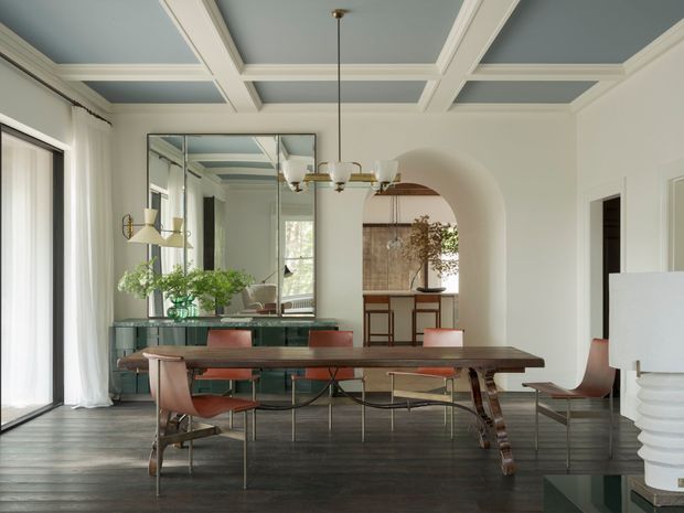 Refined dining room with a long dark wood dining table and warm-toned modern chairs, framed by a coffered blue-grey ceiling, white walls, full-height curtains, and a large mirrored wall. An arched opening leads to a compact kitchen or bar area, while a sculptural white lamp and glass console add decorative detail.