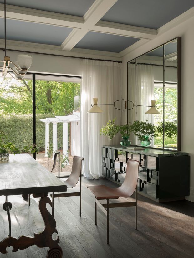 Elegant dining room with a dark timber dining table and sculptural brown leather dining chairs. The room features large black-framed glass doors opening to a leafy garden, sheer white curtains, a mirrored geometric sideboard, green glass vessels, and a coffered ceiling painted in a deep blue-grey tone with white moulded trim. A linear pendant light hangs above the table.