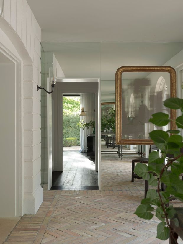 A refined interior passage or living area with a full-height mirrored wall, white-painted masonry and trim, a large antique-style gilt-framed mirror, and a warm reclaimed brick herringbone floor. The space opens toward a bright garden-facing room with dining furniture, greenery, and pendant lighting. Traditional architectural detailing is combined with reflective surfaces and restrained contemporary furnishings.