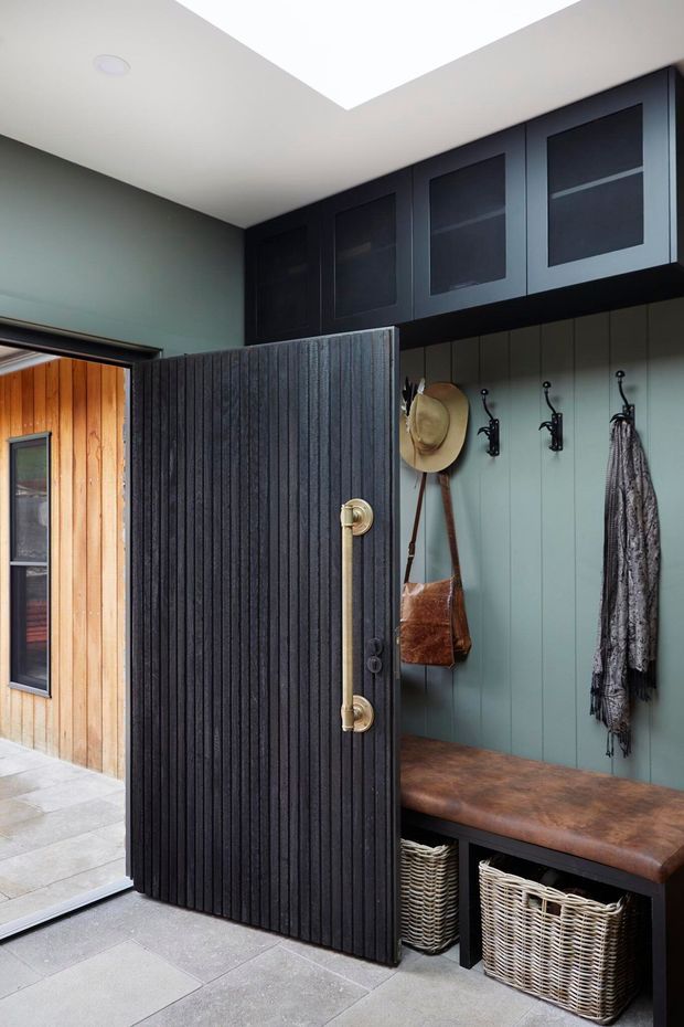 A compact, carefully fitted entryway with a dark vertically slatted pivot door, built-in overhead cupboards, wall hooks, a timber bench, and woven storage baskets. Muted green wall panelling contrasts with charcoal joinery and a warm timber-clad exterior visible through the open doorway. Large pale floor tiles and a bright ceiling lightwell add contrast.