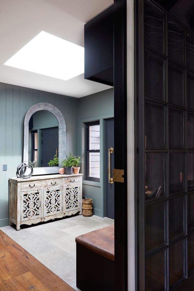 A refined residential entryway viewed through a dark paneled doorway. The space features a distressed carved console, arched wall mirror, potted greenery, blue-grey vertical wall panelling, dark trim and doors, a large skylight, and a combination of pale stone tile and warm timber flooring. The overall look blends traditional detailing with moody contemporary styling.