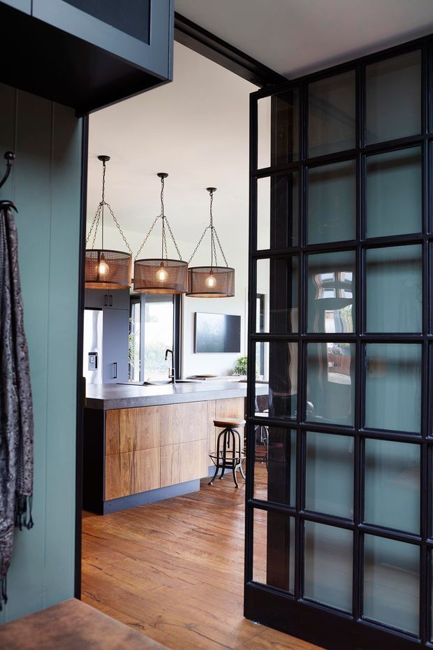 Interior view through a black-framed glazed partition door into a compact kitchen. The kitchen features a timber-front island with a light benchtop, dark cabinetry, warm timber flooring, and three pendant lights with woven metal shades. The black steel-look framed glazing creates a strong industrial architectural feature, while blue-grey wall cabinetry and frosted glass add contrast.