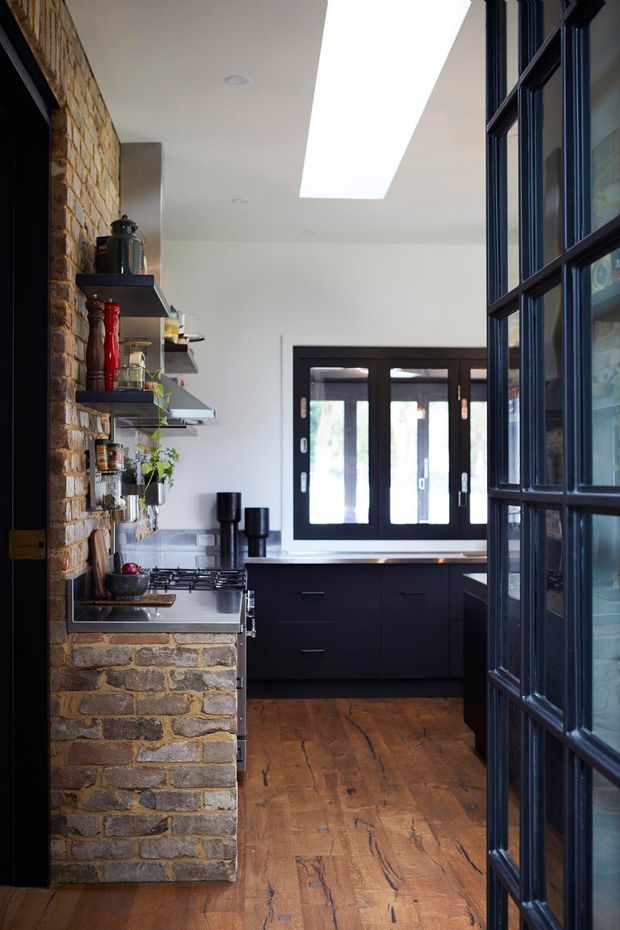 A compact, characterful kitchen viewed through a dark glazed door. The room features an exposed warm brick wall with open black shelves, dark cabinetry, a stainless-steel cooktop and rangehood, timber flooring, black-framed windows and a large ceiling skylight. The mix of exposed masonry, dark joinery and industrial-style fixtures creates a warm industrial-contemporary feel.