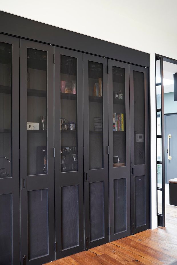 Interior view of a tall, full-height built-in storage wall with dark charcoal framed glass-panel doors and lower solid-panel cabinetry. Shelving displays books and decorative objects. The joinery stands beside a black-framed glazed opening leading toward an entry area, with warm timber flooring and a blue-grey door visible beyond.