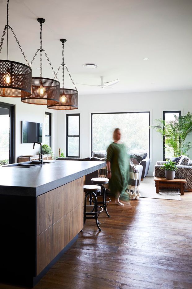 Bright open-plan interior featuring a contemporary kitchen with a large dark stone island, timber cabinetry, black-framed windows, and a cluster of perforated metal pendant lights. The island opens toward a living area with patterned armchairs, indoor palms, a wall-mounted television, and polished timber flooring.