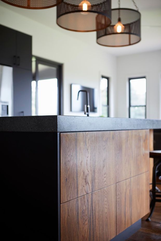 Close-up view of a contemporary kitchen island with a dark, textured benchtop and black side panels, paired with vertically grained timber cabinetry. The bright kitchen has dark-framed windows, a black tap and sink, and several suspended wire-mesh pendant lights.