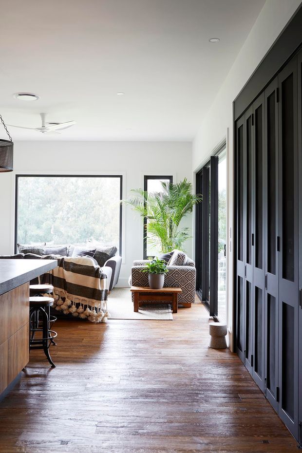 Bright open-plan interior showing a living area beside a kitchen or breakfast bar. The space has white walls and ceiling, warm timber flooring, extensive black-framed glazing and a full-height bank of dark built-in cabinetry. A sofa, patterned armchairs, indoor palm, pendant light and ceiling fan create a relaxed, contemporary atmosphere.