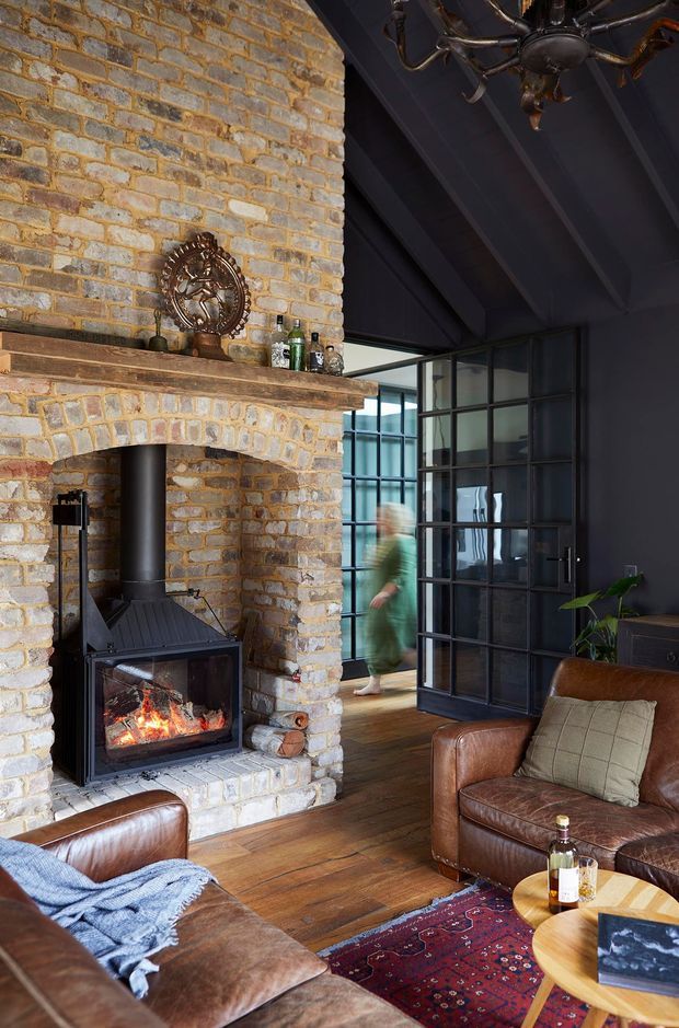 A warm, characterful living room centred on a substantial exposed brick fireplace with a modern wood-burning stove. The room has dark painted exposed roof beams, black steel-framed glazed doors, wide timber flooring, brown leather sofas, a patterned rug, and a round timber coffee table. A glimpse of an adjoining space is visible through the open steel-framed doors.