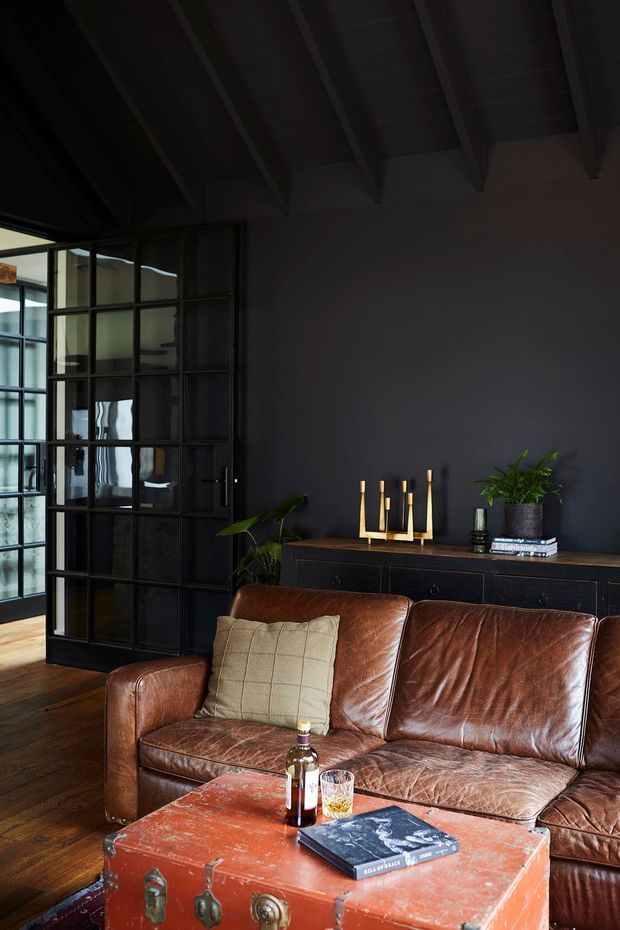 A moody, refined living room with a large worn brown leather sofa, a distressed red-orange trunk used as a coffee table, and a dark timber sideboard. The room features a charcoal painted wall, black steel-framed glazed doors, exposed dark ceiling beams, warm timber flooring, decorative candles, and indoor plants.