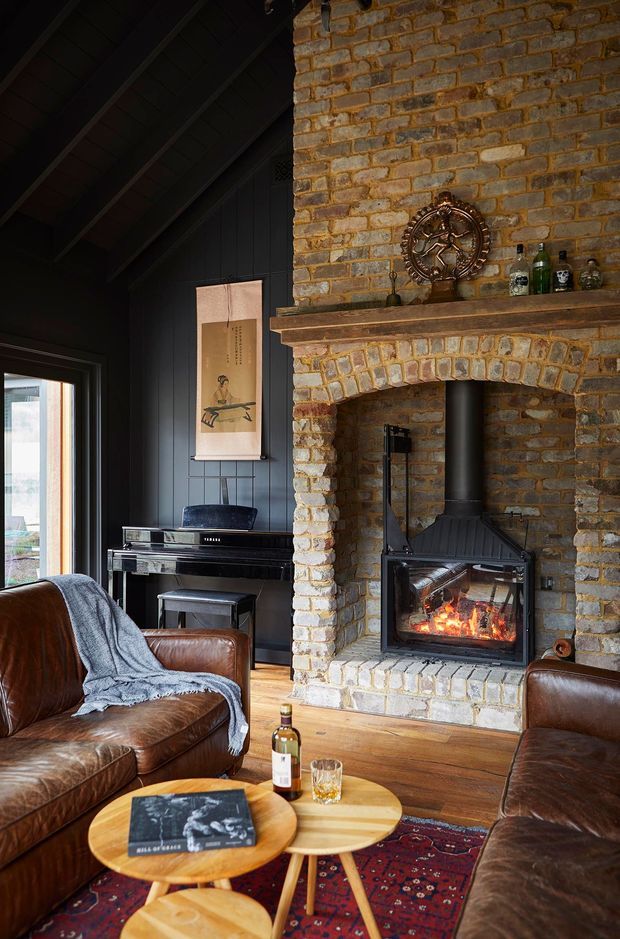 A cozy, dark-toned living room centred on a substantial exposed brick fireplace with a lit wood-burning stove. The room features black-painted vertical wall panelling and a dark pitched ceiling, warm timber flooring, a black upright piano, and vintage brown leather sofas and armchair. Layered round timber coffee tables, a patterned rug, artwork, bottles, and a draped throw add a relaxed, eclectic character.