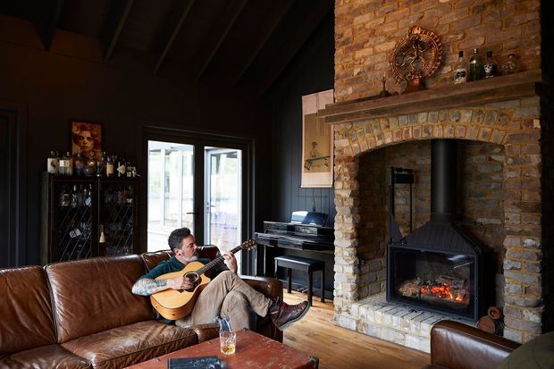 A warm, rustic living room with dark painted walls, exposed timber ceiling beams, wide timber flooring, and a prominent rough brick masonry fireplace with a wood-burning stove. A leather sofa, piano, drinks cabinet, artwork, and a person playing guitar create a relaxed, intimate atmosphere. Glazed French doors bring in daylight.