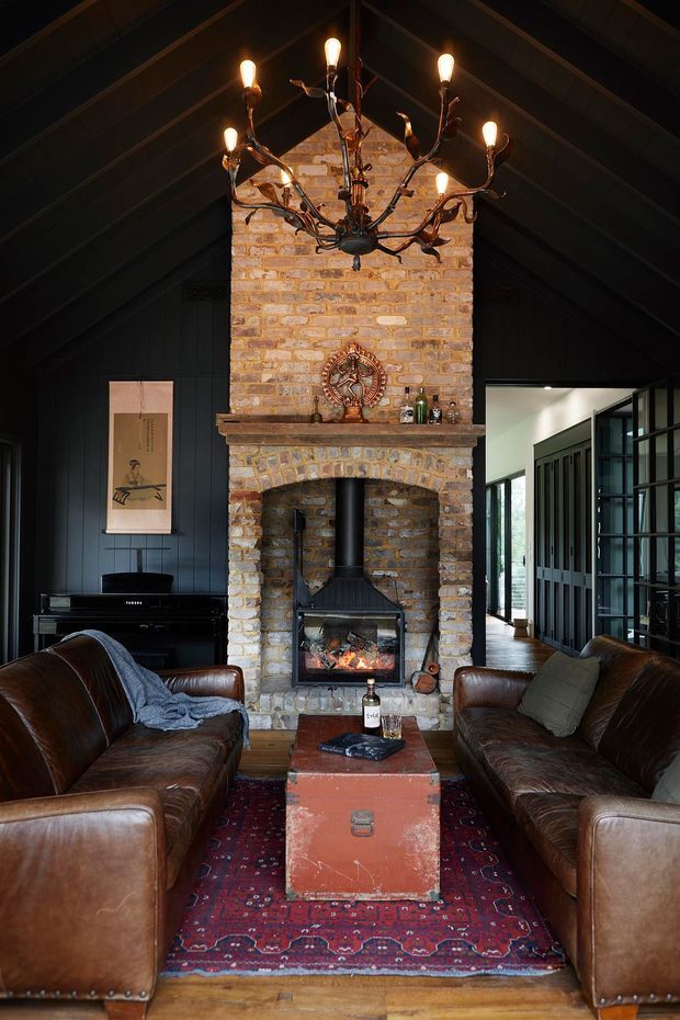 A cozy, dramatic living room centred on a large exposed-brick fireplace with a wood-burning stove. Dark vertical wall panelling and a pitched, timber-lined ceiling frame the room, while two worn brown leather sofas face each other across a patterned rug and vintage trunk coffee table. A sculptural rustic chandelier hangs overhead, with a piano and glazed partition visible to the sides.