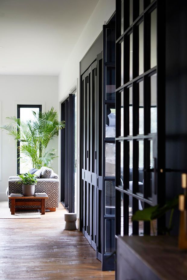 A bright, refined interior hallway with warm timber flooring, white walls, and a dramatic series of black-framed glazed and paneled doors or screens along one side. The hallway leads toward a relaxed seating nook with a patterned armchair, timber coffee table, potted palm, and large black-framed window. Reflections reveal additional furnishings beyond the glazing.