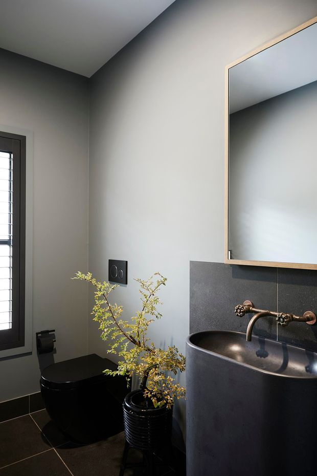 A compact, refined bathroom with dark tiled flooring, charcoal walls and a matching sculptural basin vanity. A black toilet sits beside a tall louvred window, while a large timber-framed mirror and aged bronze wall-mounted tapware add warmth and contrast. A potted branch provides a natural decorative accent.