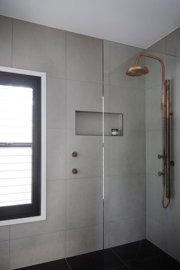 Modern walk-in shower with large grey wall tiles, a frameless glass shower screen, black tiled floor, recessed niche, and distinctive copper-toned exposed shower fittings. A louvred window with a dark frame provides natural light.