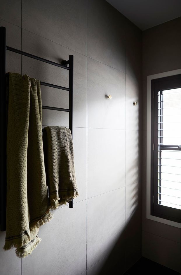 A moody contemporary bathroom corner with large pale grey tiled wall panels, a matte black ladder-style heated towel rail holding olive and beige towels, dark flooring, and a narrow shuttered window admitting strong natural light.