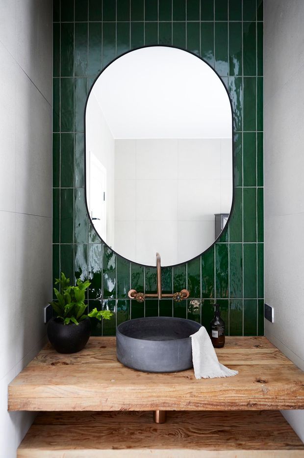 Compact bathroom vanity with a large arched, black-framed mirror set against glossy dark green vertical tiles. The space features a rustic solid timber benchtop and open timber shelf, a round dark stone vessel basin, copper-toned wall-mounted tapware, a matte black planter, and white tiled side walls. The mirror reflects a bright tiled shower or bathroom area.