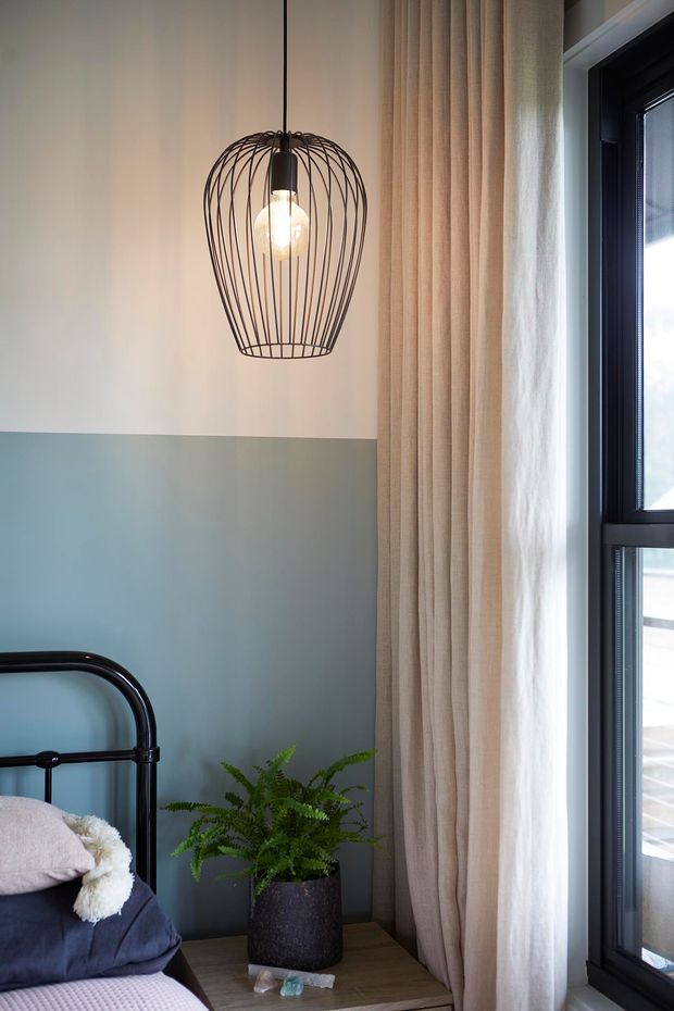 A softly lit bedroom corner with a black metal bed frame, layered dark and pale bedding, a two-tone blue-grey and off-white wall, a hanging black wire-cage pendant light, full-height beige curtains, and a large black-framed window. A potted fern sits on a timber bedside table, creating a calm contemporary interior.