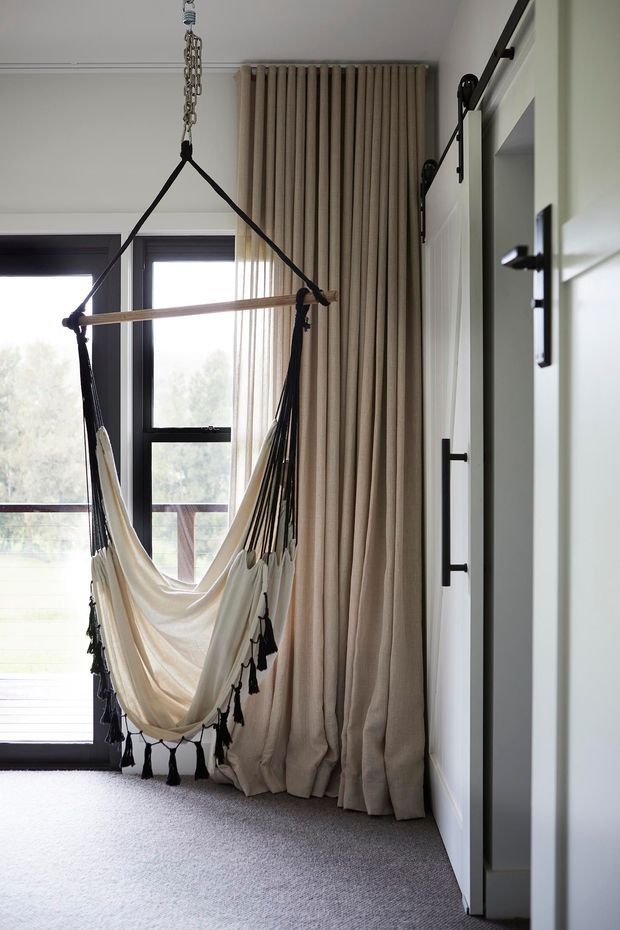 A bright, compact interior corner with a cream fabric hanging hammock suspended in front of a large black-framed glazed door or window. Full-height beige curtains frame the glazing, while white paneled sliding barn-style doors with black hardware line the right side. The room has a light grey carpet, white walls, and a relaxed, softly textured aesthetic.