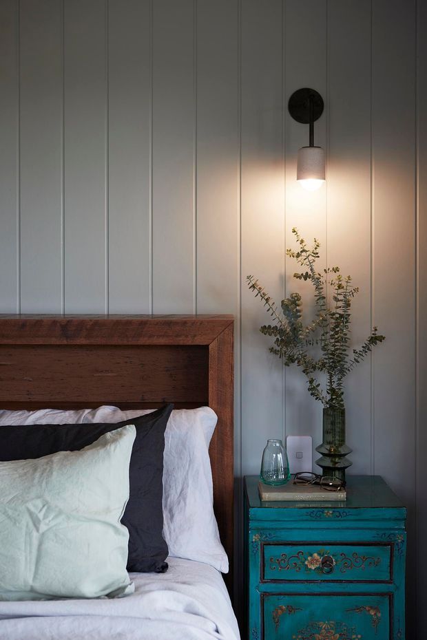 A moody bedroom vignette featuring a timber bed with white and charcoal bedding, a vertically panelled pale wall, and a richly painted turquoise bedside chest with decorative detailing. A warm pendant wall light illuminates a glass vase of eucalyptus beside the bed.