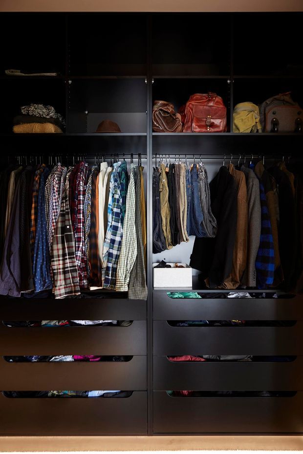 A dark, bespoke walk-in wardrobe with floor-to-ceiling black or charcoal cabinetry. The upper open shelves display handbags and hats, while hanging rails hold a broad collection of shirts, jackets, trousers and coats. Multiple deep drawers provide folded-clothing storage, with integrated low-level lighting creating a dramatic, refined atmosphere.