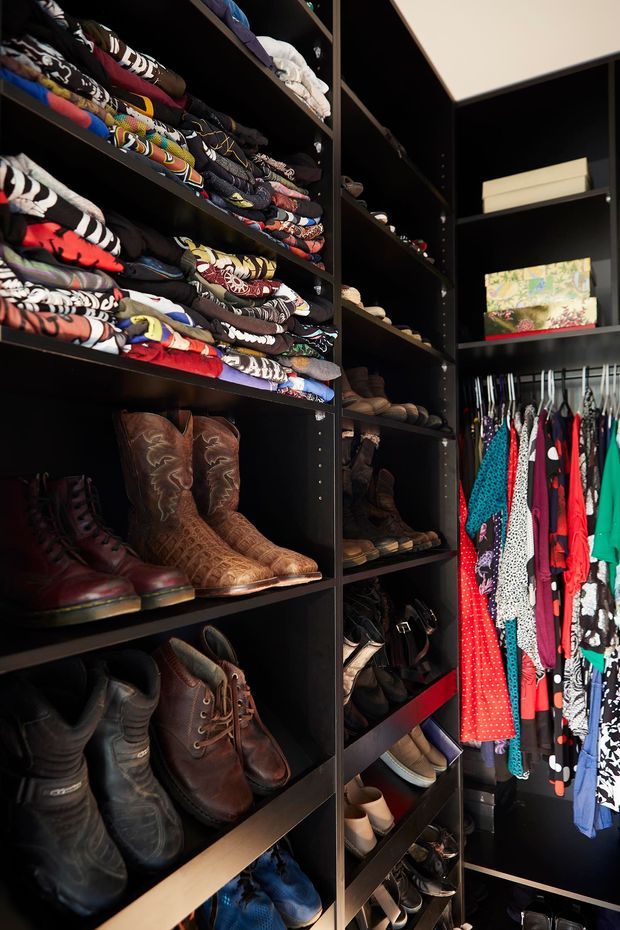 A compact walk-in wardrobe or dressing room with floor-to-ceiling black built-in shelving and hanging rails. Folded colourful clothing fills the upper shelves, while boots, shoes and accessories occupy lower compartments. Bright garments hang on the right, creating a bold contrast against the dark cabinetry.