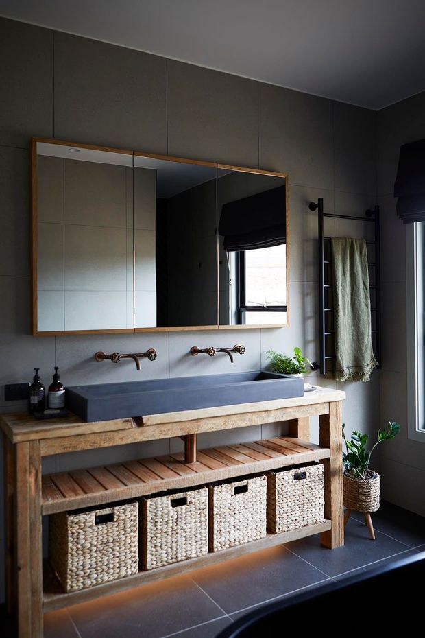 A moody, rustic bathroom vanity with a large framed mirrored cabinet, twin wall-mounted brass taps, a deep rectangular vessel sink, and a substantial reclaimed timber console with open shelving and woven storage baskets. Dark large-format tiled walls and floor, black-framed windows, a towel ladder, and potted greenery create a warm, textural interior.