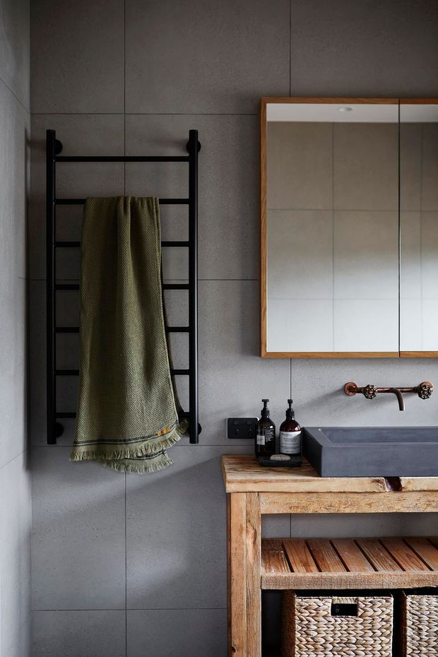 A moody, modern bathroom vignette featuring large grey wall tiles, a black ladder-style heated towel rail, and a timber-framed mirrored cabinet above a dark rectangular basin. A rustic timber vanity with an open slatted shelf and woven baskets sits beneath the basin, complemented by copper-toned wall-mounted tapware and dark accessories.