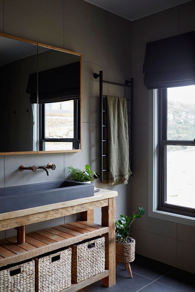 A moody, rustic bathroom featuring a freestanding timber vanity with a pale stone benchtop, dark rectangular vessel basin, and aged copper wall-mounted tap. A large timber-framed mirror, black metal towel ladder, woven textile, roller blinds, dark wall panels, tiled flooring, woven vanity baskets, and indoor plants complete the space.