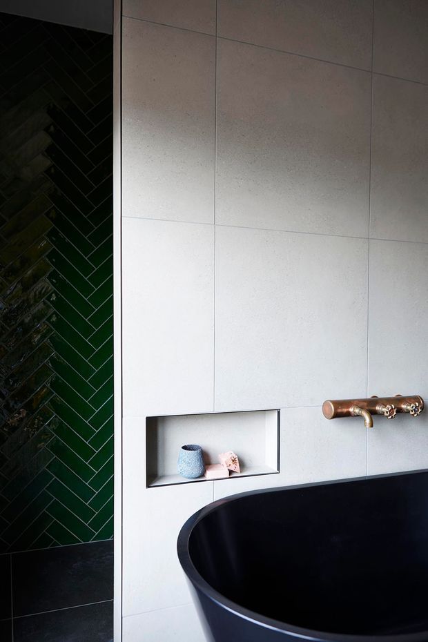 Close-up view of a contemporary bathroom featuring a deep black freestanding bathtub, large pale grey wall tiles, a dark green herringbone-tiled shower wall, a recessed wall niche, and a copper wall-mounted bath mixer. The image has a refined, moody palette with strong contrast between the light tiled wall and dark fixtures.