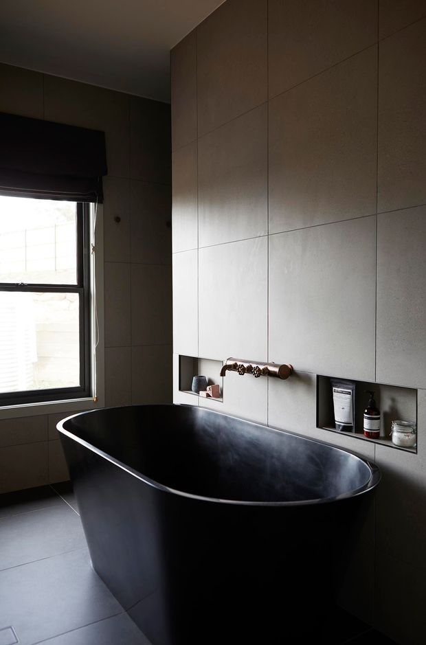 A moody contemporary bathroom featuring a freestanding matte-black bathtub, large-format grey wall and floor tiles, dark window framing, and recessed wall niches with bath products. A wall-mounted aged brass tap and fittings provide a warm metallic accent, while natural light enters through the window.