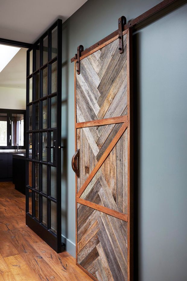Interior view featuring a prominent rustic reclaimed-wood barn-style sliding door with herringbone and chevron geometric paneling, dark metal track hardware, and a curved metal handle. The door is set against a muted blue-grey wall, beside black-framed industrial-style glass partitions. Warm timber flooring and a glimpse of a dark kitchen or living area are visible beyond.