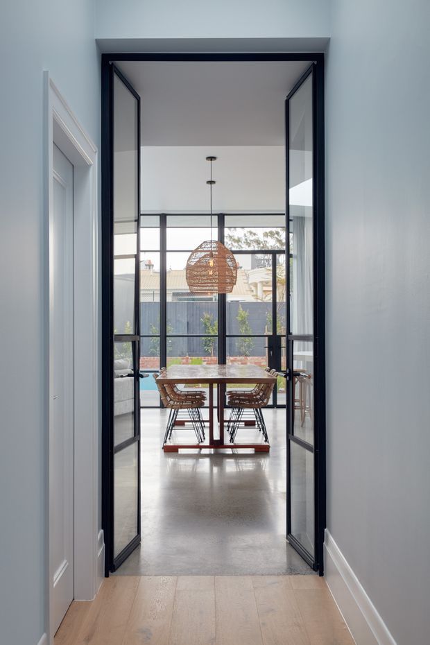 A bright interior view through a hallway into a dining area. Black-framed glazed double doors open onto a compact dining space with a wood-toned rectangular table and woven chairs, backed by large black-framed windows overlooking a fenced outdoor area. A woven pendant light hangs above the table. The foreground hallway has pale blue-grey walls, white trim, and light timber flooring, while the dining area has a polished concrete floor.