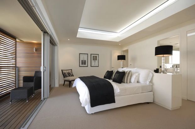 Bright, refined contemporary bedroom with a large upholstered bed dressed in white linens and layered black, grey, and striped cushions. The room has soft beige carpeting, white walls and built-in cabinetry, black pendant lamps, framed artwork, and a full-width skylight or recessed ceiling feature. Large sliding glazed doors open to a timber-lined balcony or outdoor sitting area with horizontal timber blinds.