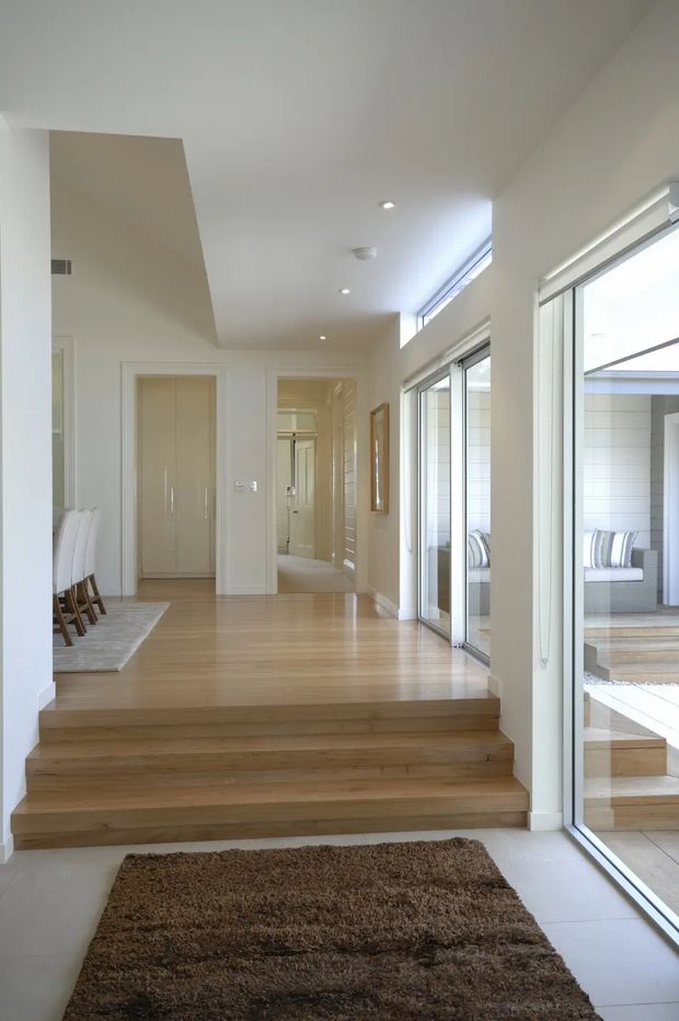 Bright, refined residential interior showing a short set of timber steps leading into an open hallway and dining area. The space has pale timber flooring, white walls and ceiling, recessed downlights, tall windows, clerestory glazing, and large sliding glass doors opening to a timber deck. A built-in wardrobe and corridor are visible beyond, with dining chairs on the left and reflected outdoor seating through the glazing.