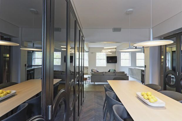 A refined open-plan interior viewed from a dining area, with a long light-wood dining table, upholstered chairs, pendant lighting, and decorative dark metal-and-glass room dividers. A contemporary living room with sectional seating and a wall-mounted television is visible beyond, while a kitchen or bar area appears to the right. The space combines warm timber tones, dark framed glazing and partitions, pale walls, and patterned timber flooring.