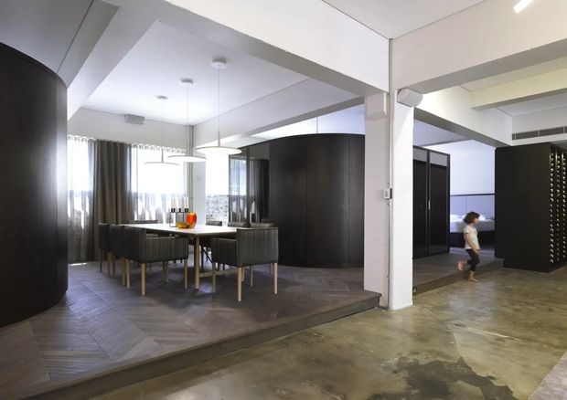 Open-plan contemporary interior featuring a raised dining area with a dark timber dining table and upholstered chairs, surrounded by curved dark joinery and room dividers. Herringbone timber flooring defines the dining zone, while the adjacent lower floor is polished concrete. White structural columns and beams, pendant lighting, and full-height curtains contribute to the modern, minimalist composition.