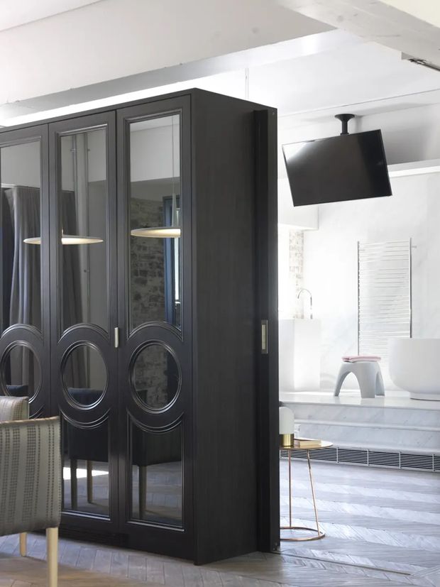A sophisticated open-plan bathroom viewed behind a tall, dark timber partition or wardrobe with decorative mirrored panels and oval mouldings. The bright bathroom includes a freestanding white bathtub, wall-mounted towel rail, pale stone or tiled surfaces, a suspended black television, and herringbone flooring. A partial upholstered chair and small side table are visible in the foreground. The overall composition is monochromatic, high-contrast, and refined, combining dark joinery with a bright contemporary bathroom.